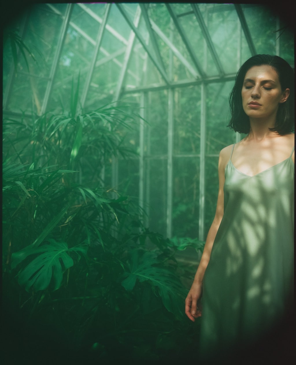 A dark-haired woman in a pale green slip dress stands in an overgrown glasshouse, dappled light across her shoulders.